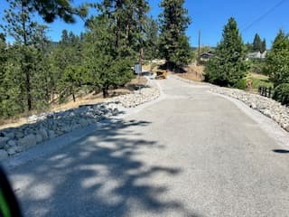 Paved road lined with large rocks winding through a forest with construction equipment ahead.