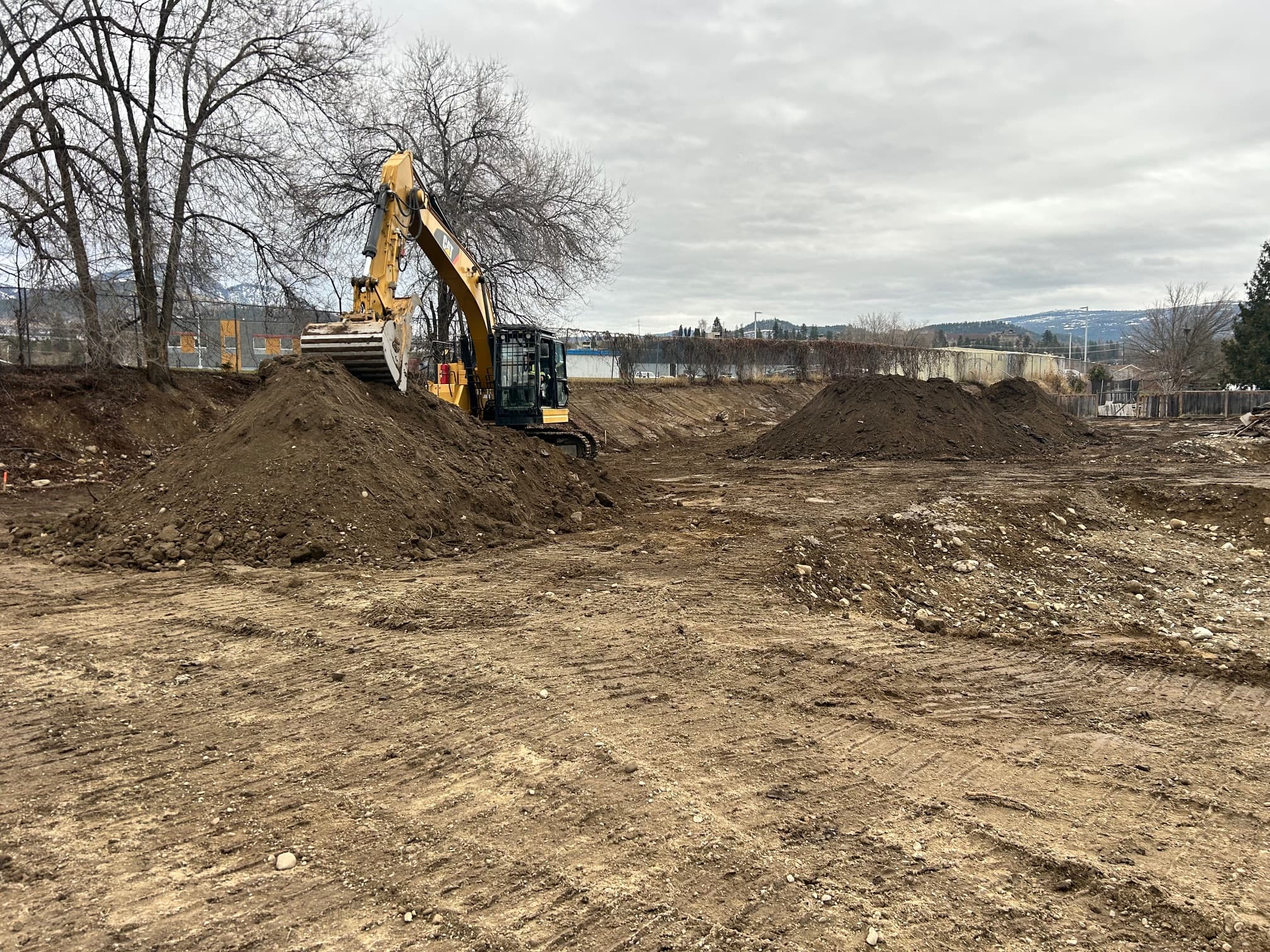 Yellow excavator atop a large mound of dirt at a construction site under cloudy skies.