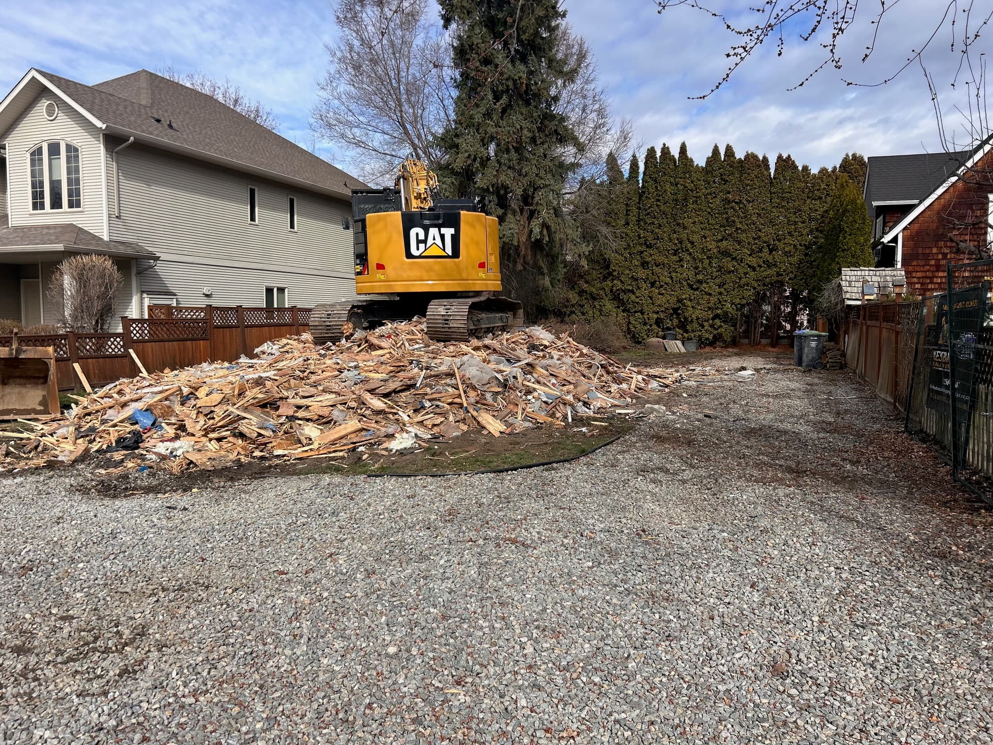 Yellow CAT excavator sits on a large pile of wooden debris in a residential yard.